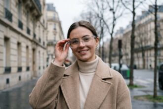 Jeune femme parisienne portant des lunettes transparentes tendance hiver 2026 sur un trottoir haussmannien